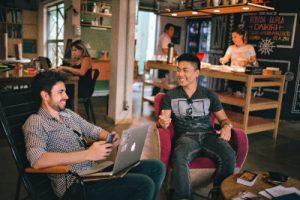 A stock photo of two men smiling and talking to each other in a coffee shop while one works on a laptop, illustrating the principle of internal networking.