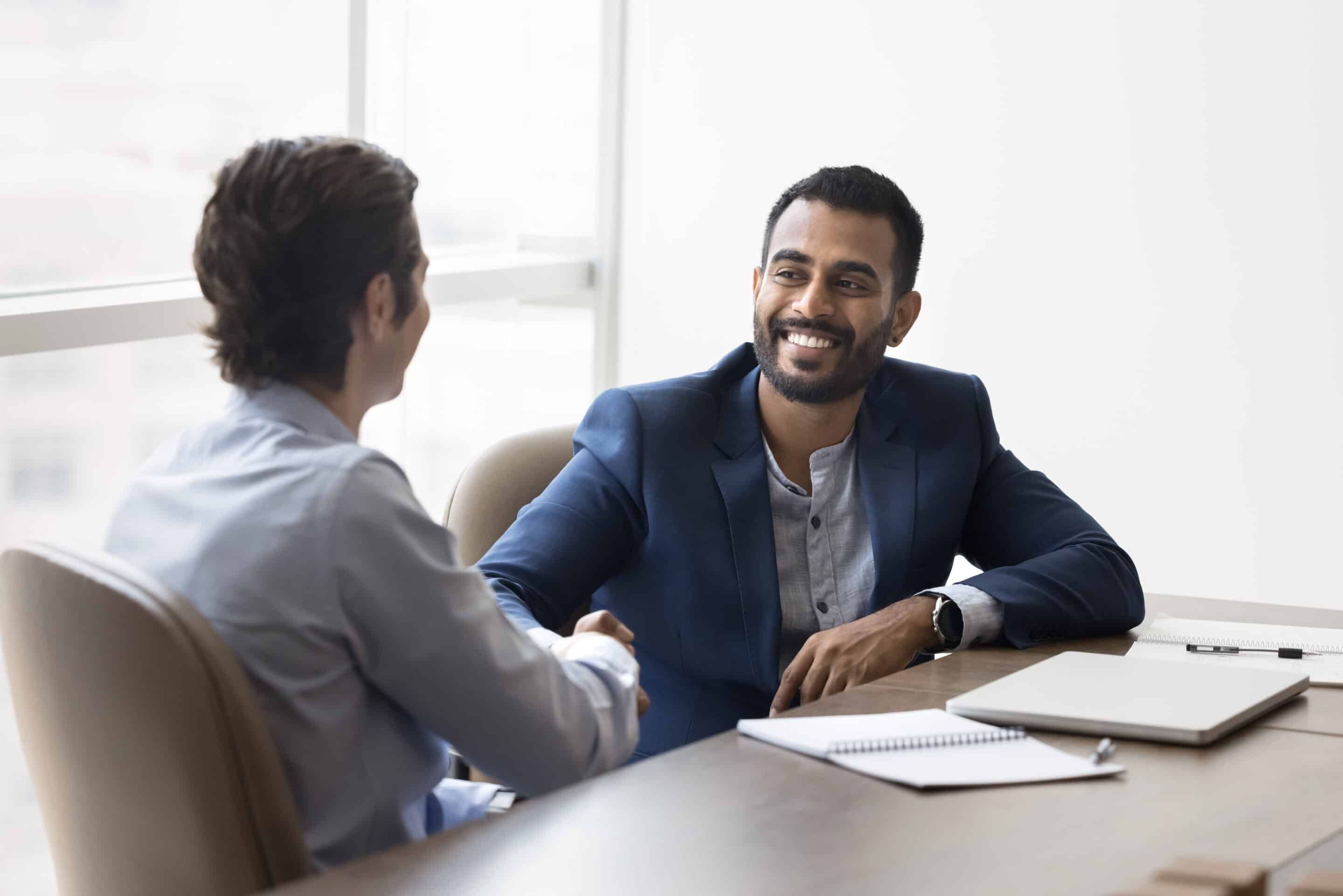 Happy young Indian business owner man shaking hands with male partner at meeting table, hiring new employee after interview, thanking financial advisor for consultation, starting partnership